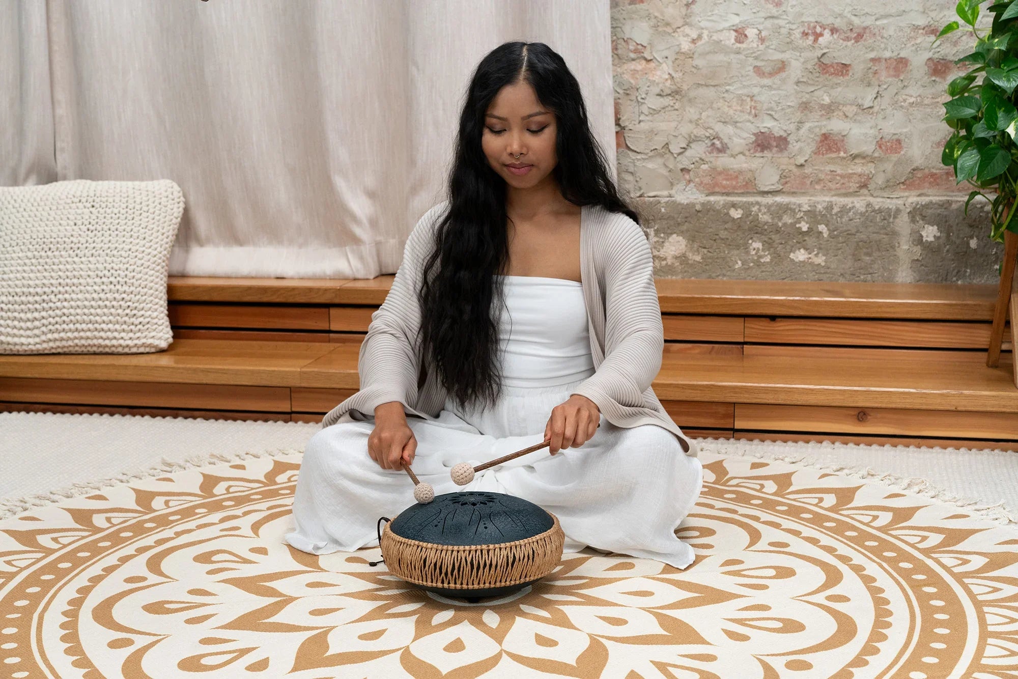 Woman playing a steel tongue drum for sound healing on a mandala patterned rug indoors.