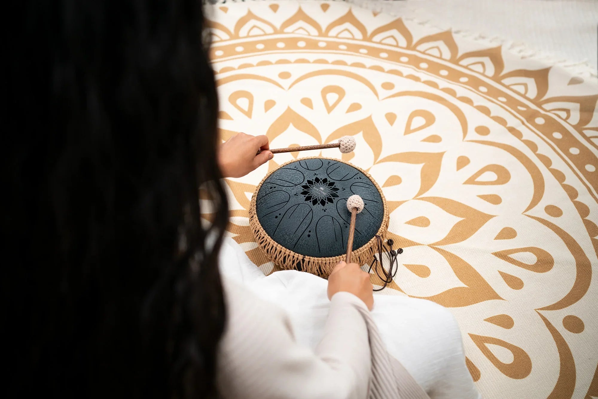 Person playing black steel tongue drum with mallets on a mandala mat, sound healing instrument