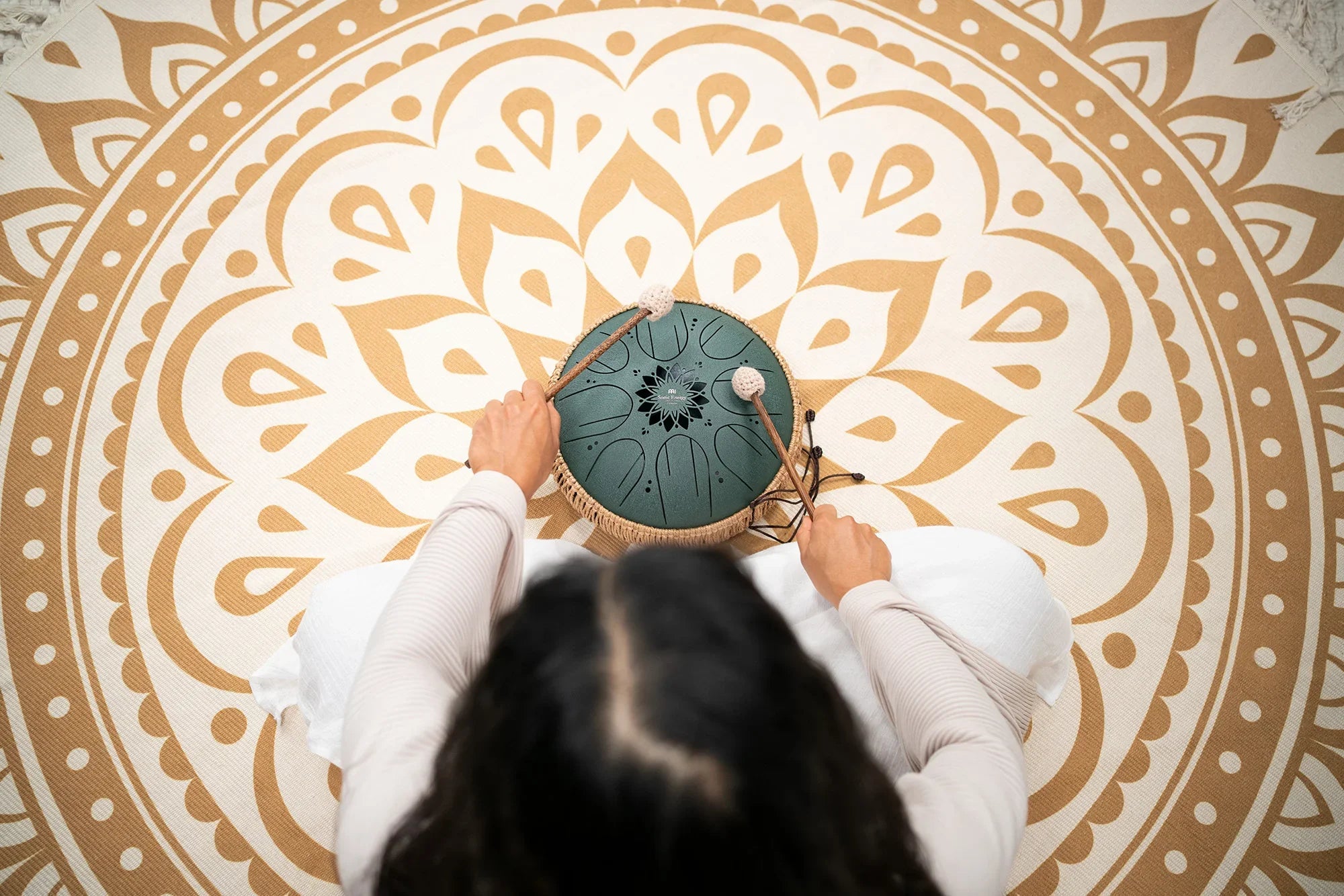 Person playing a green steel tongue drum with mallets on a large mandala mat
