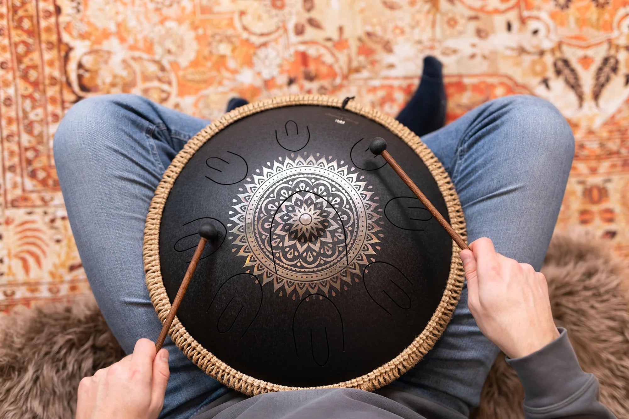 Person playing a black tongue drum with mandala design, using mallets, indoors on patterned rug