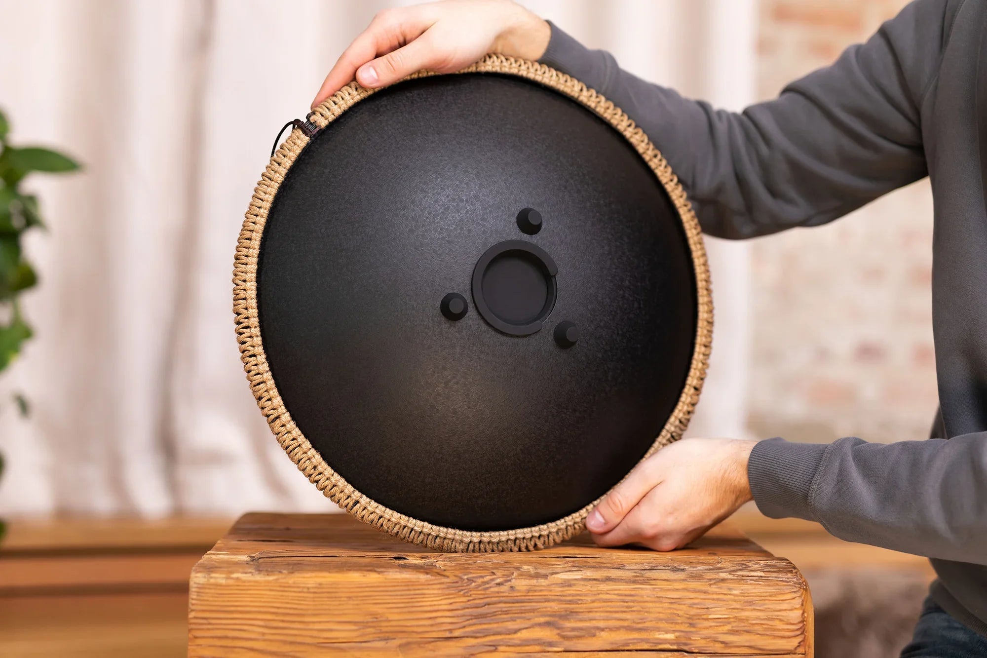 Person holding black handpan drum with woven rim on wooden table, energy sound healing instrument