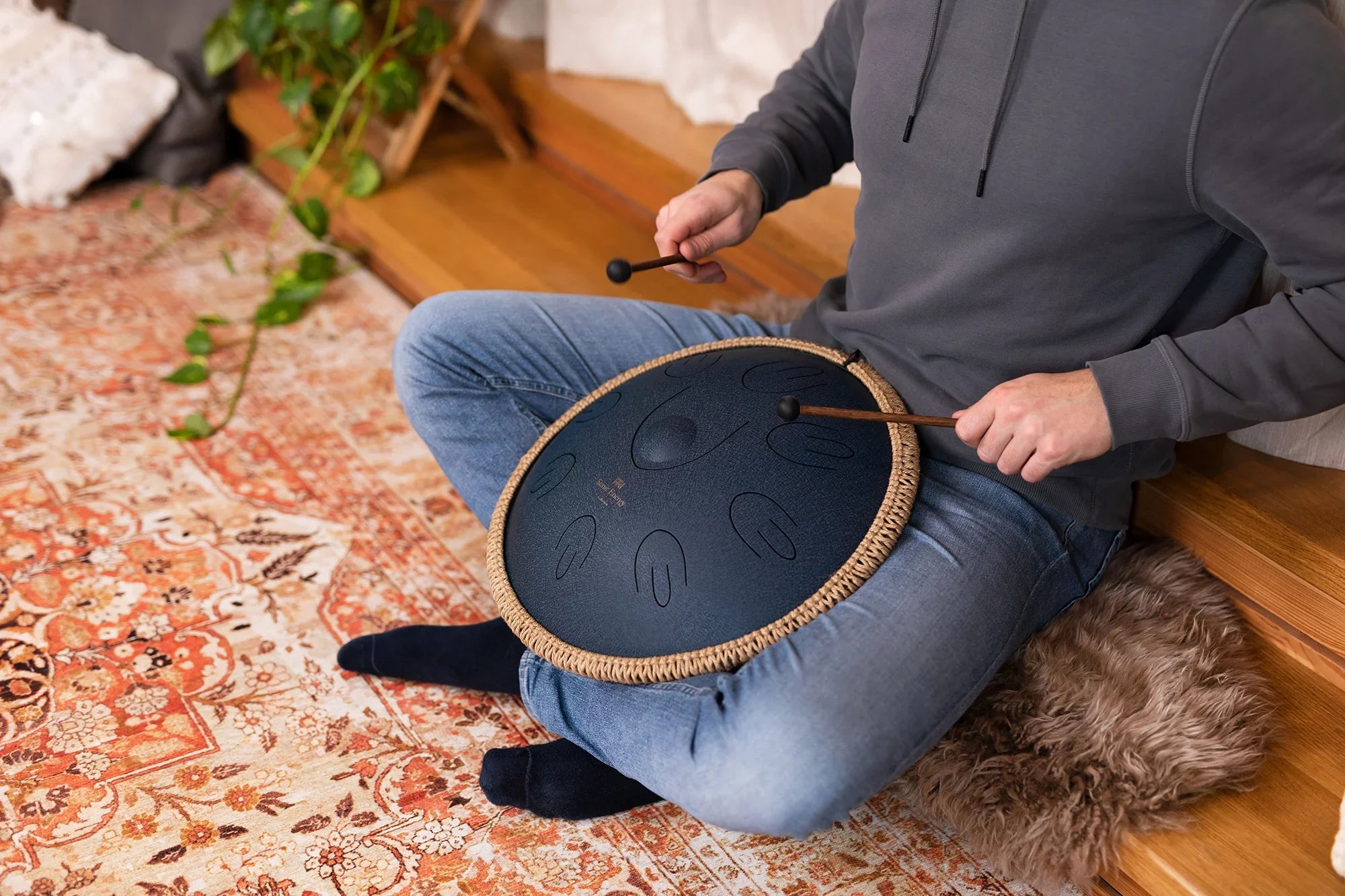 Person playing blue steel tongue drum with mallets in cozy indoor setting