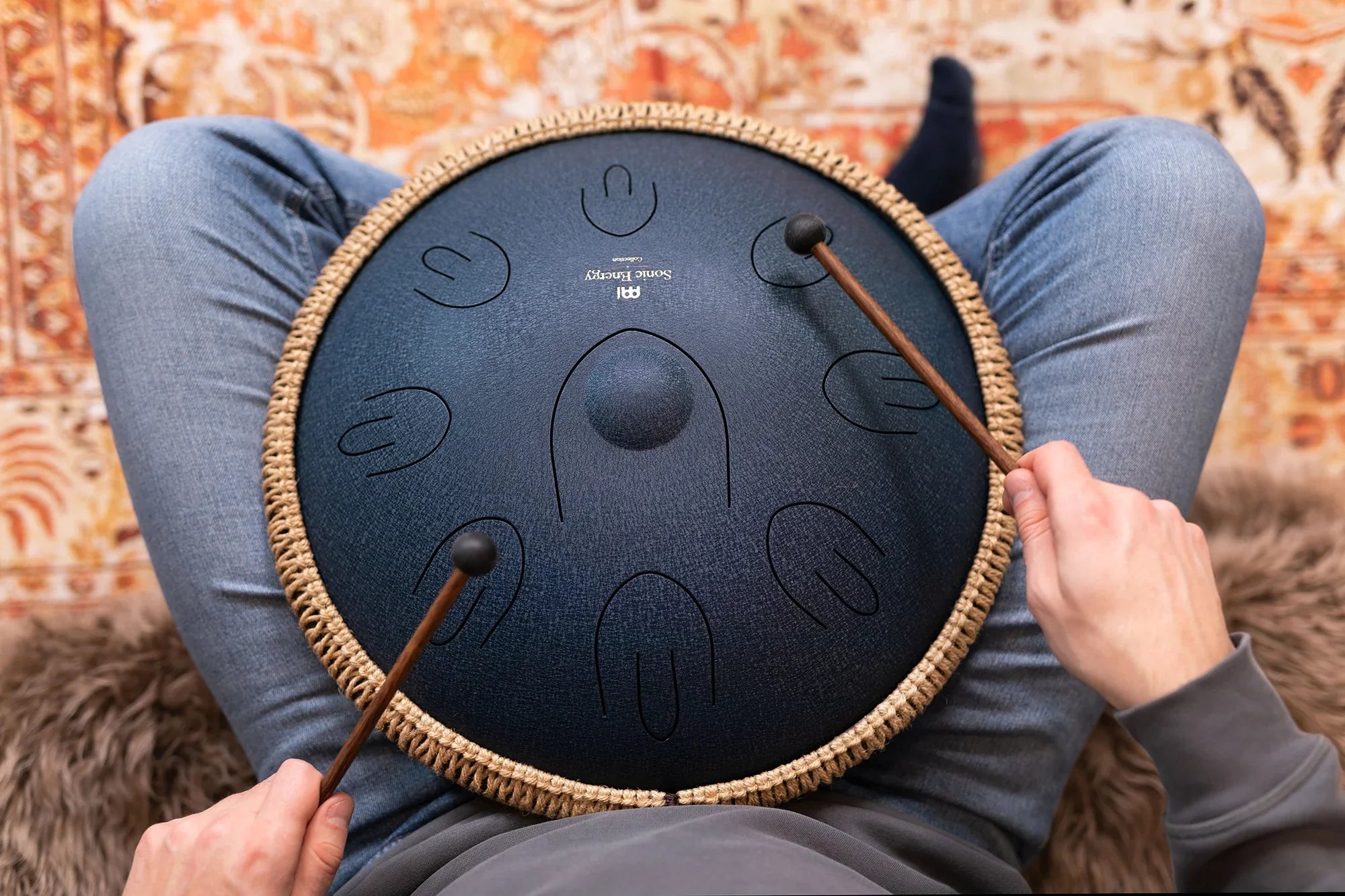 Person playing a steel tongue drum with mallets, sitting on the floor