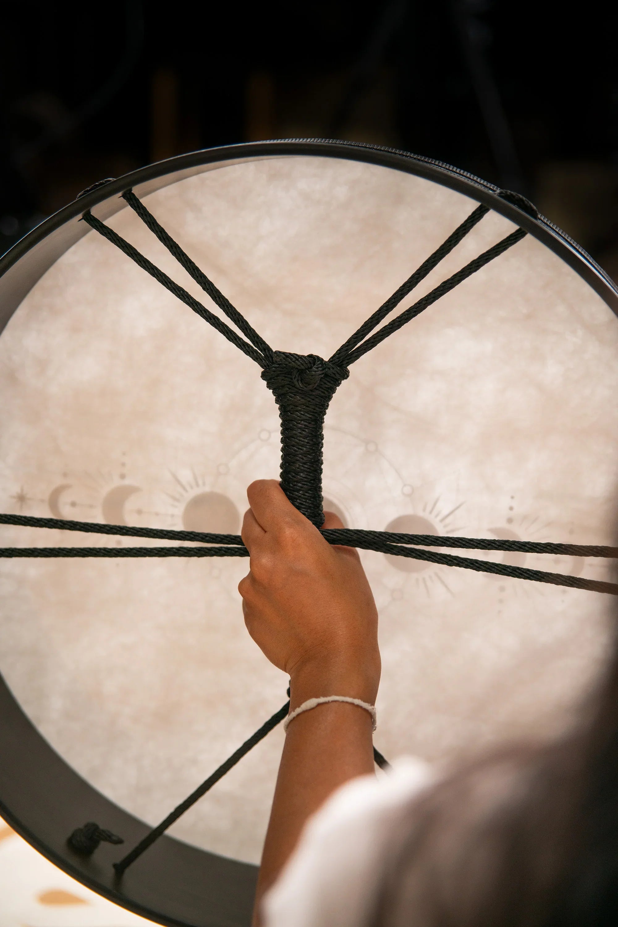 Hand holding rope handle of a healing frame drum with moon phase symbols.