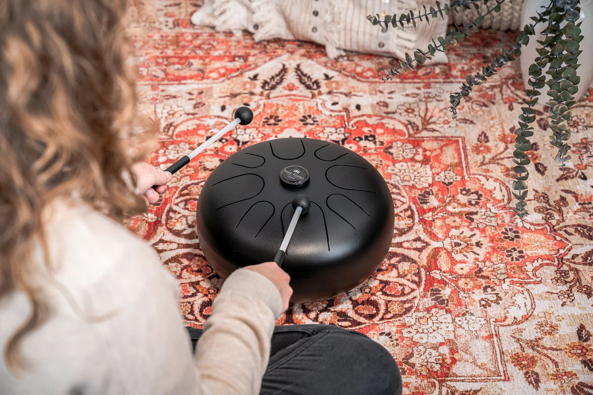 Person playing a black steel tongue drum with mallets on a patterned rug