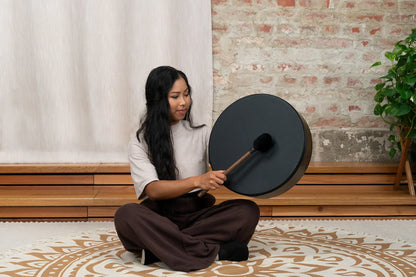 Woman playing a black frame drum for sound healing in a peaceful indoor setting.