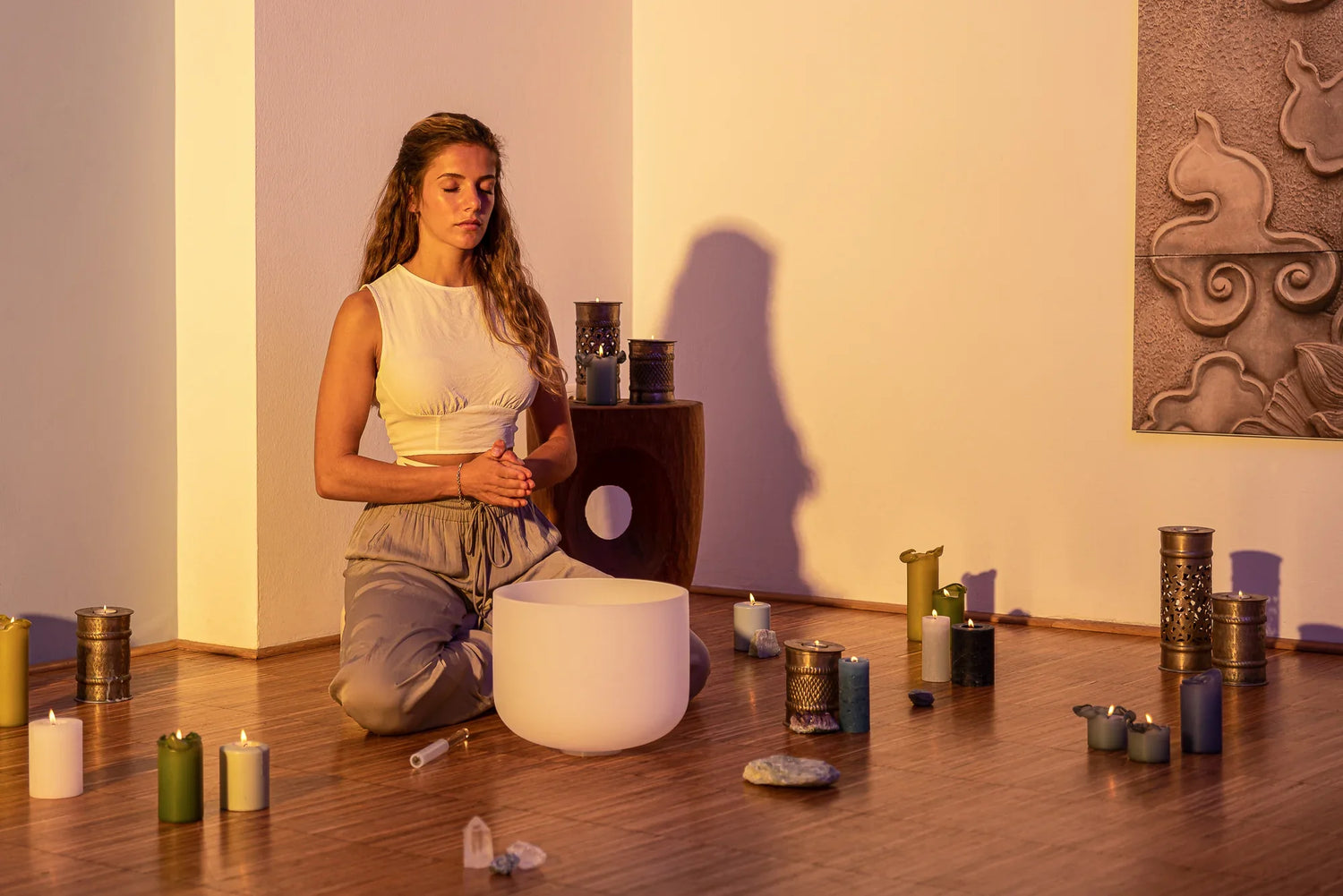 Woman meditating with a white crystal singing bowl, surrounded by candles and crystals on a wooden floor