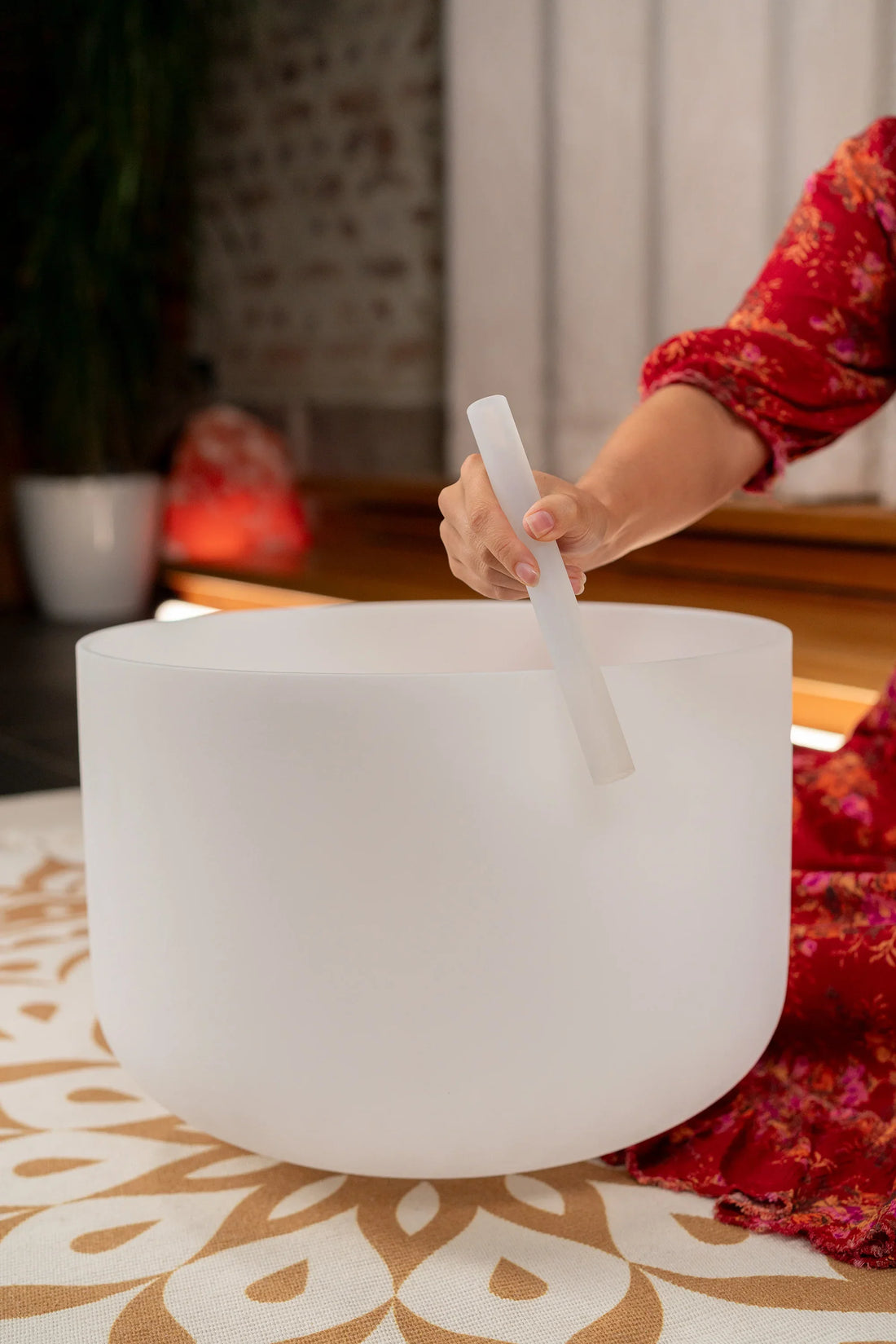 Hand playing white crystal singing bowl with mallet on patterned mat, sound healing instrument