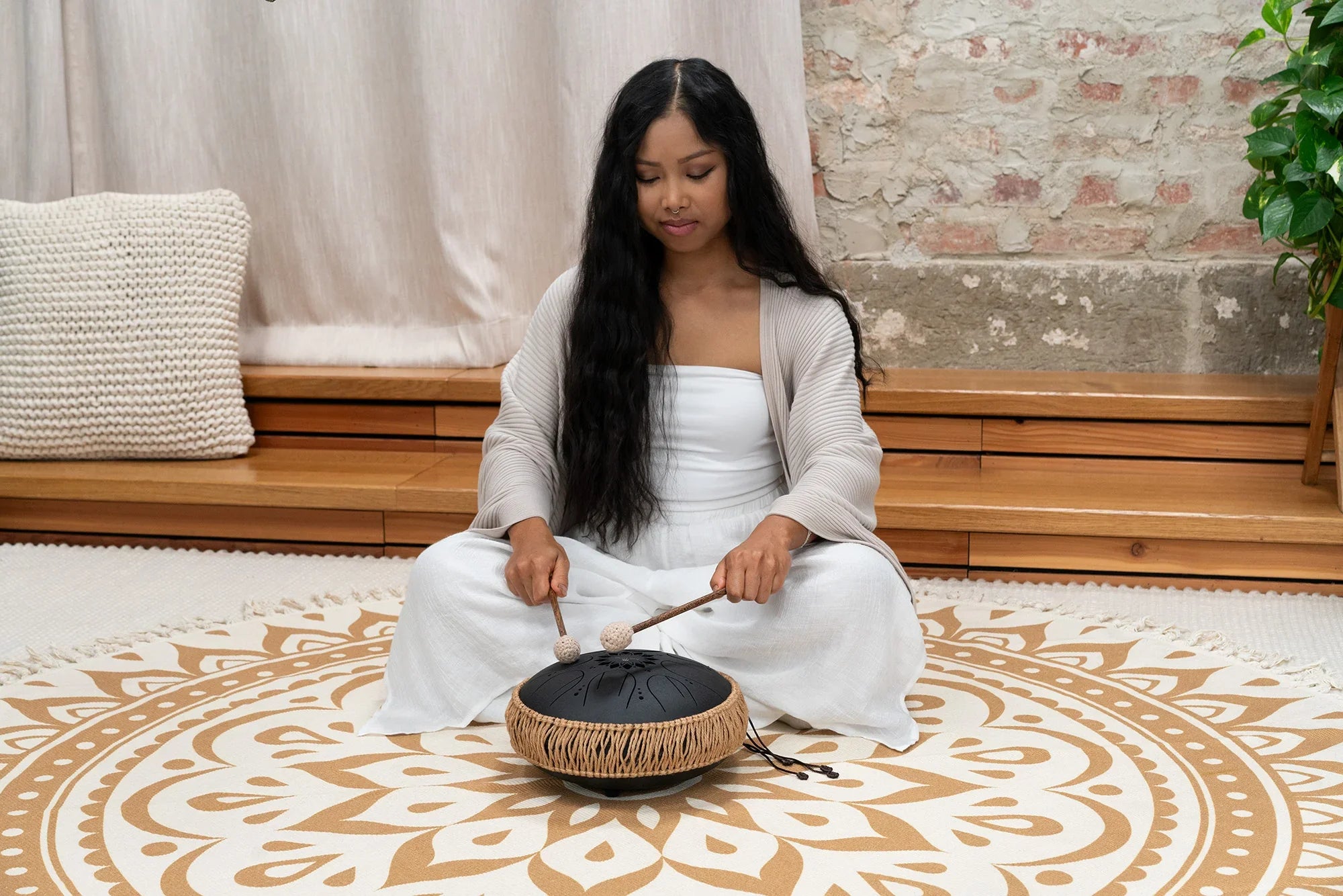 Woman playing a black steel tongue drum with mallets in a peaceful meditation space