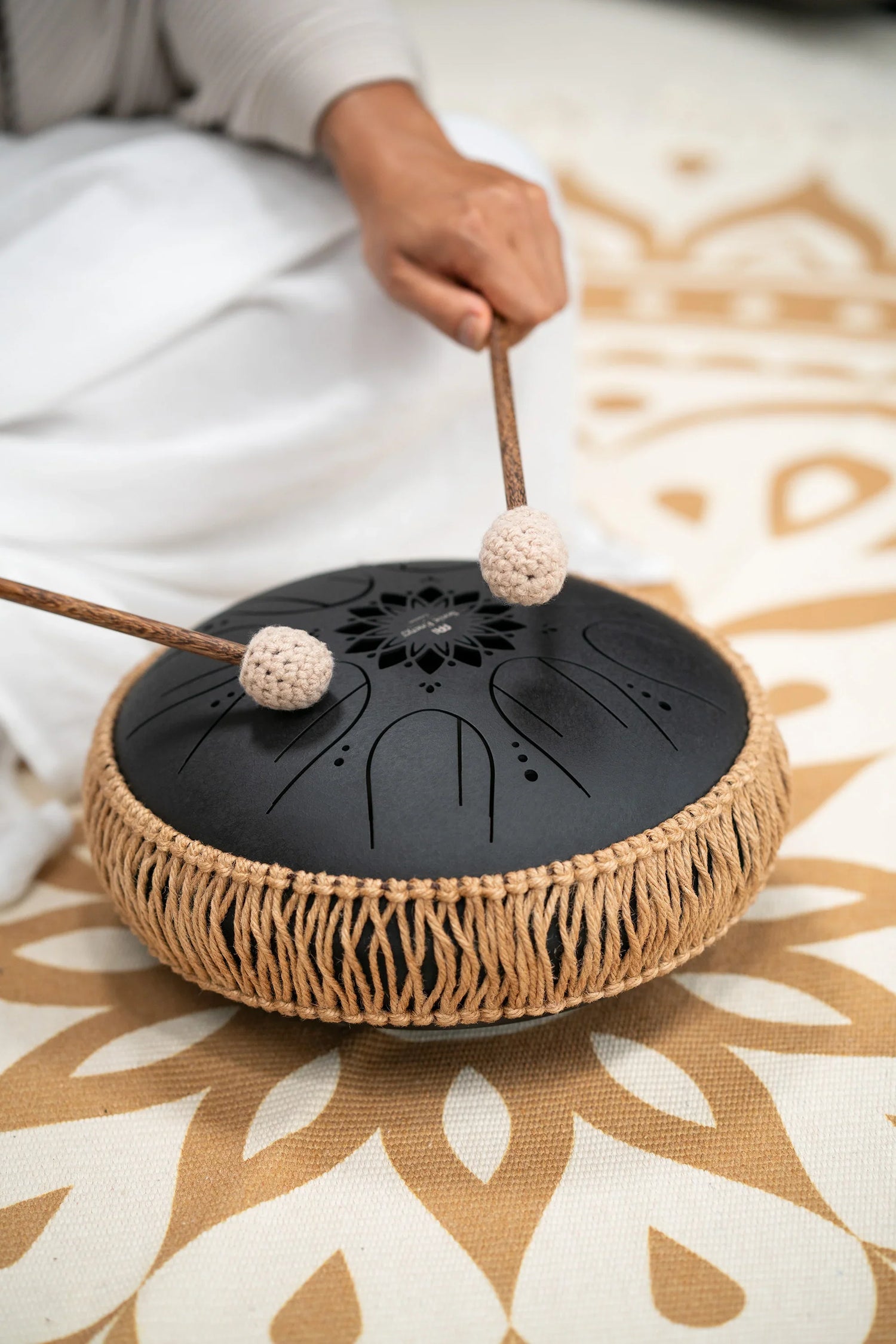 Hand playing a black steel tongue drum with mallets on a patterned mat, sound healing instrument