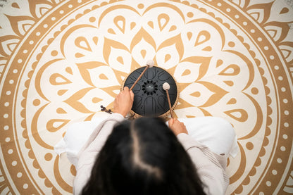 Person playing a black steel tongue drum with mallets on a mandala mat