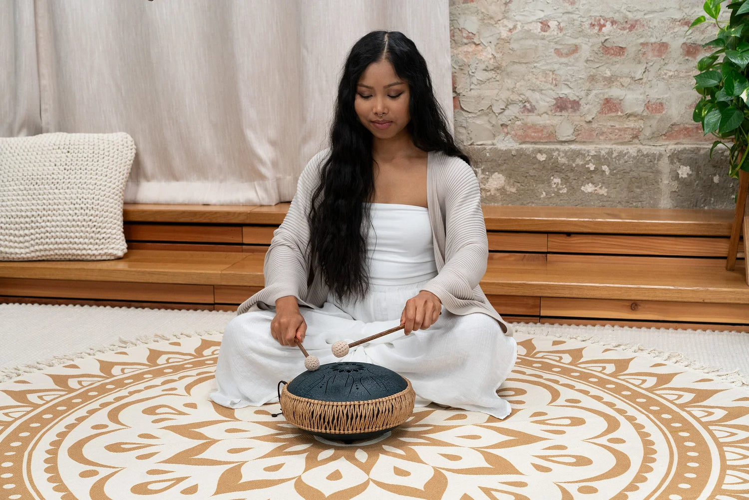 Woman playing a steel tongue drum for sound healing on a mandala patterned rug indoors.