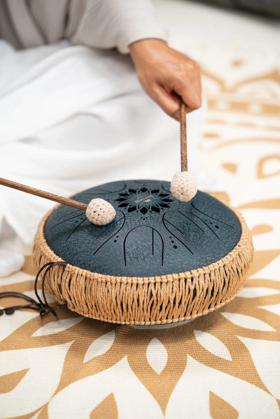Hand playing black steel tongue drum with mallets on patterned mat, for sound healing or meditation