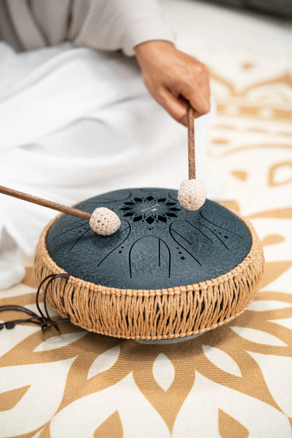 Hand playing black steel tongue drum with mallets on patterned mat, for sound healing or meditation