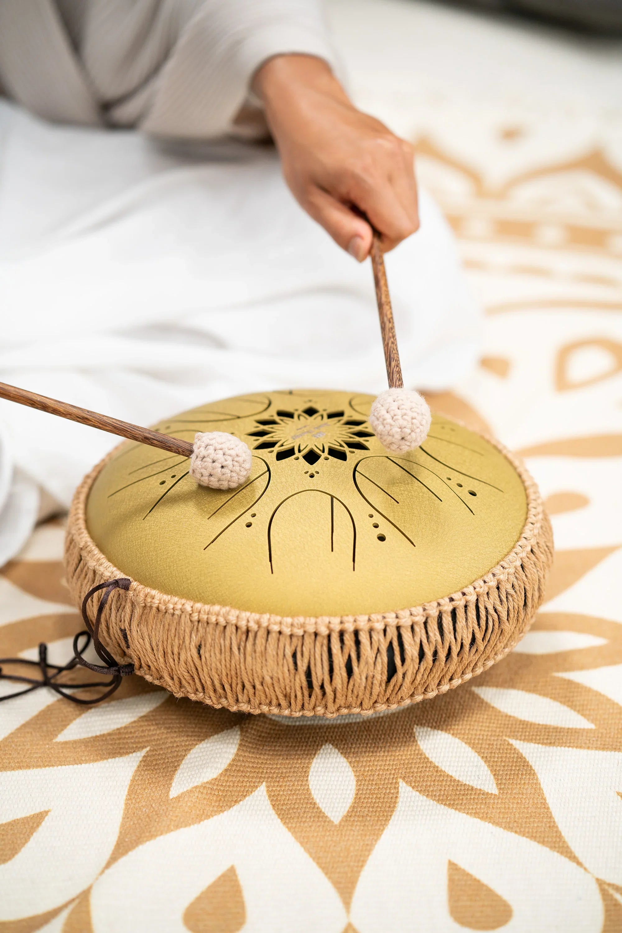 Hand playing gold steel tongue drum with mallets on patterned mat, sound healing instrument