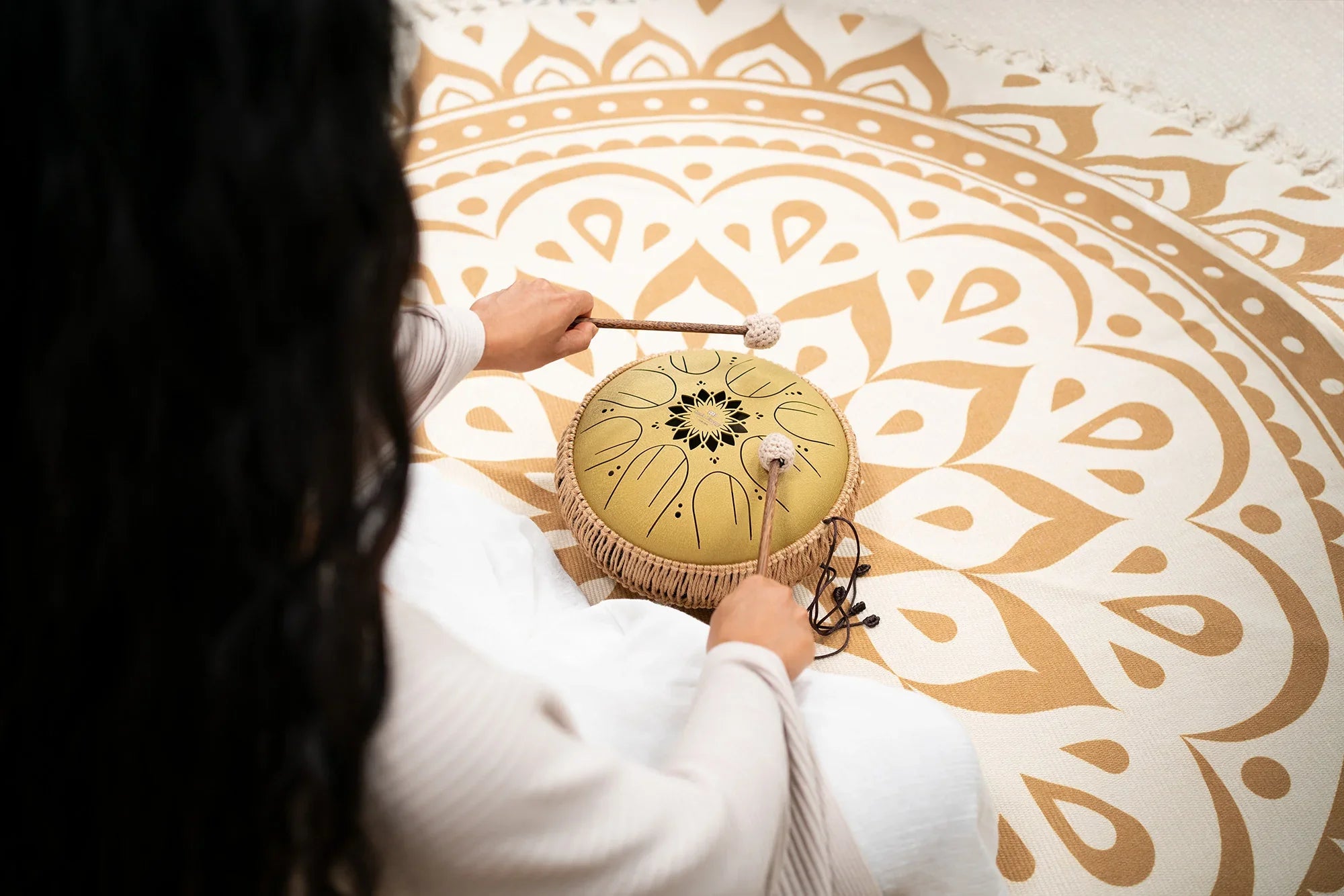 Person playing a handpan drum with mallets on a mandala-patterned rug, sound healing instrument