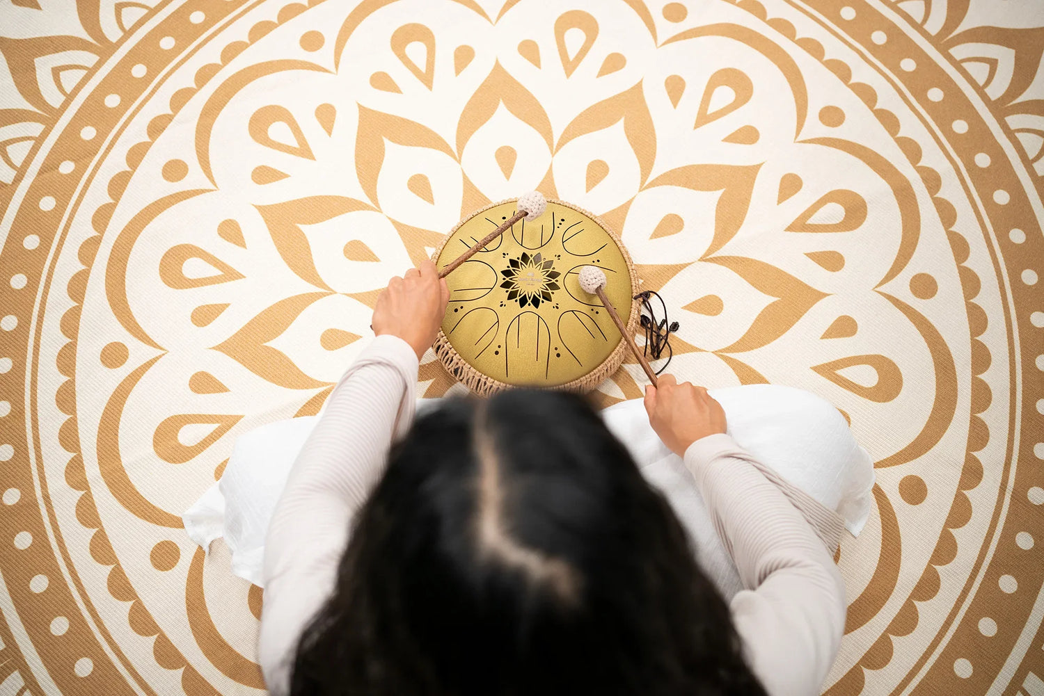 Person playing a steel tongue drum with mallets on a mandala mat for sound healing.