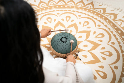 Person playing a green steel tongue drum with mallets on a decorative mandala mat