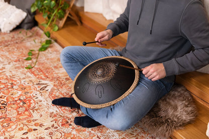 Person playing a black steel tongue drum with mallets in a cozy indoor setting