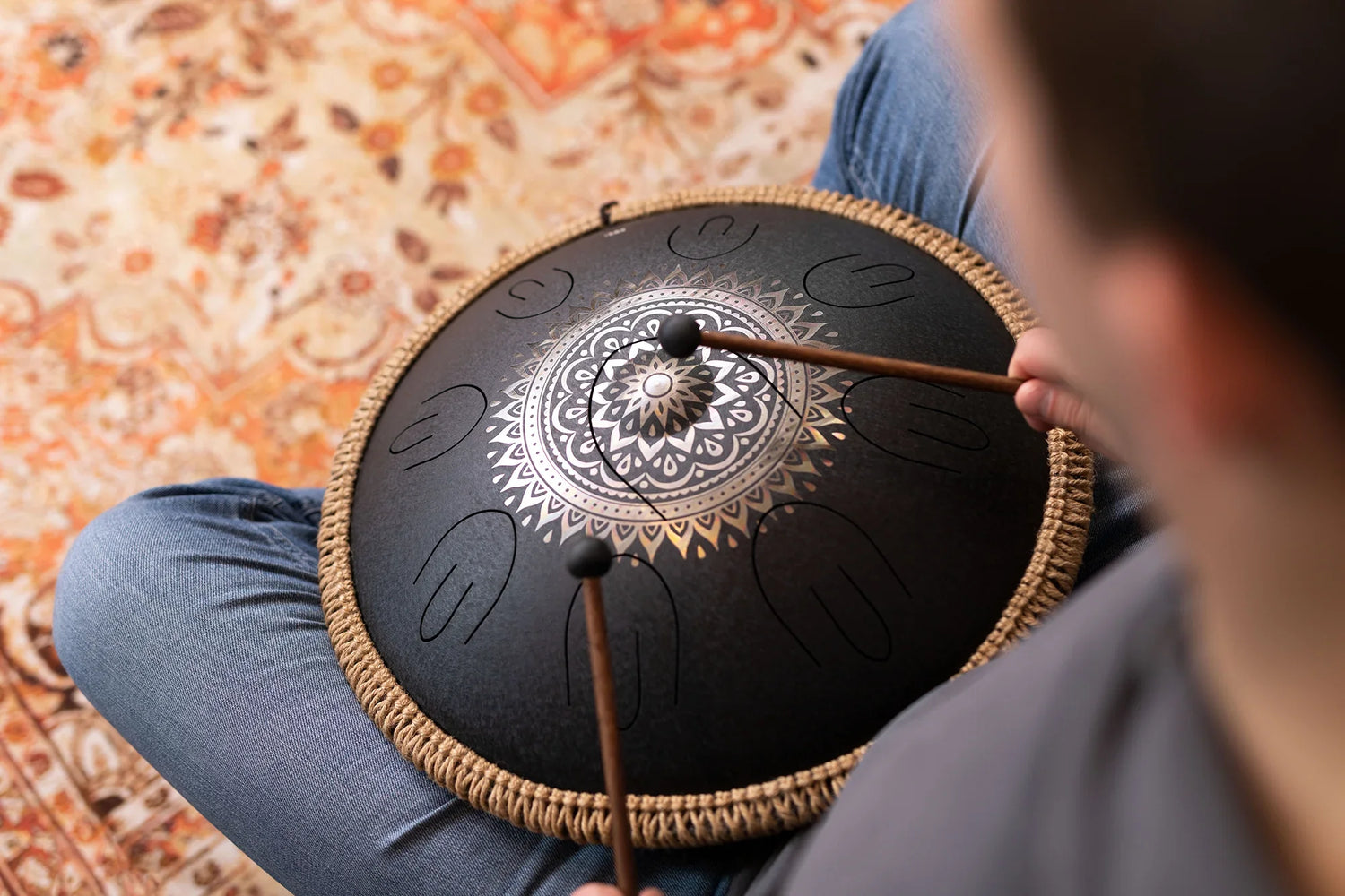 Person playing a black steel tongue drum with mallets, seated on patterned carpet