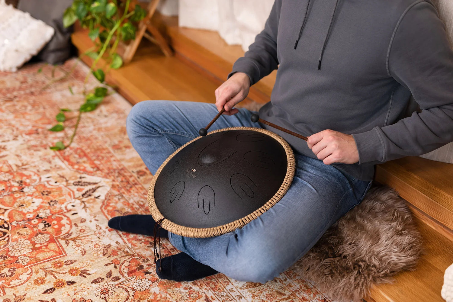 Person playing a black tongue drum with mallets in a cozy indoor setting on a patterned rug.