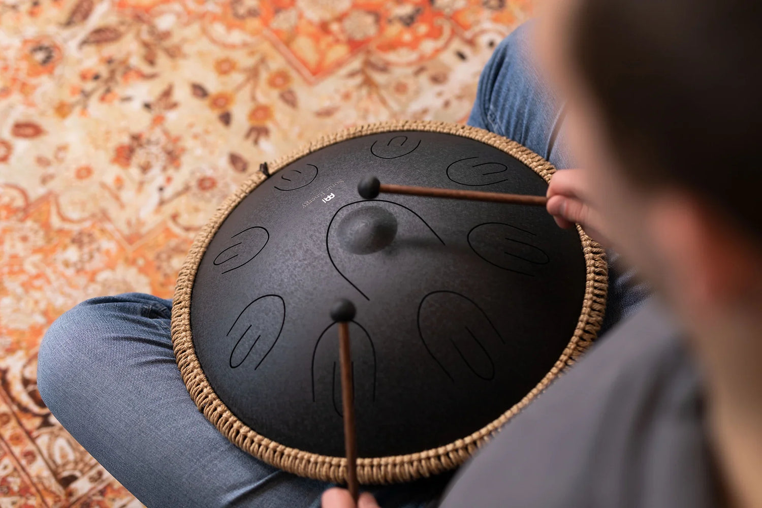 Person playing black tongue drum with mallets on patterned rug, sound healing instrument.