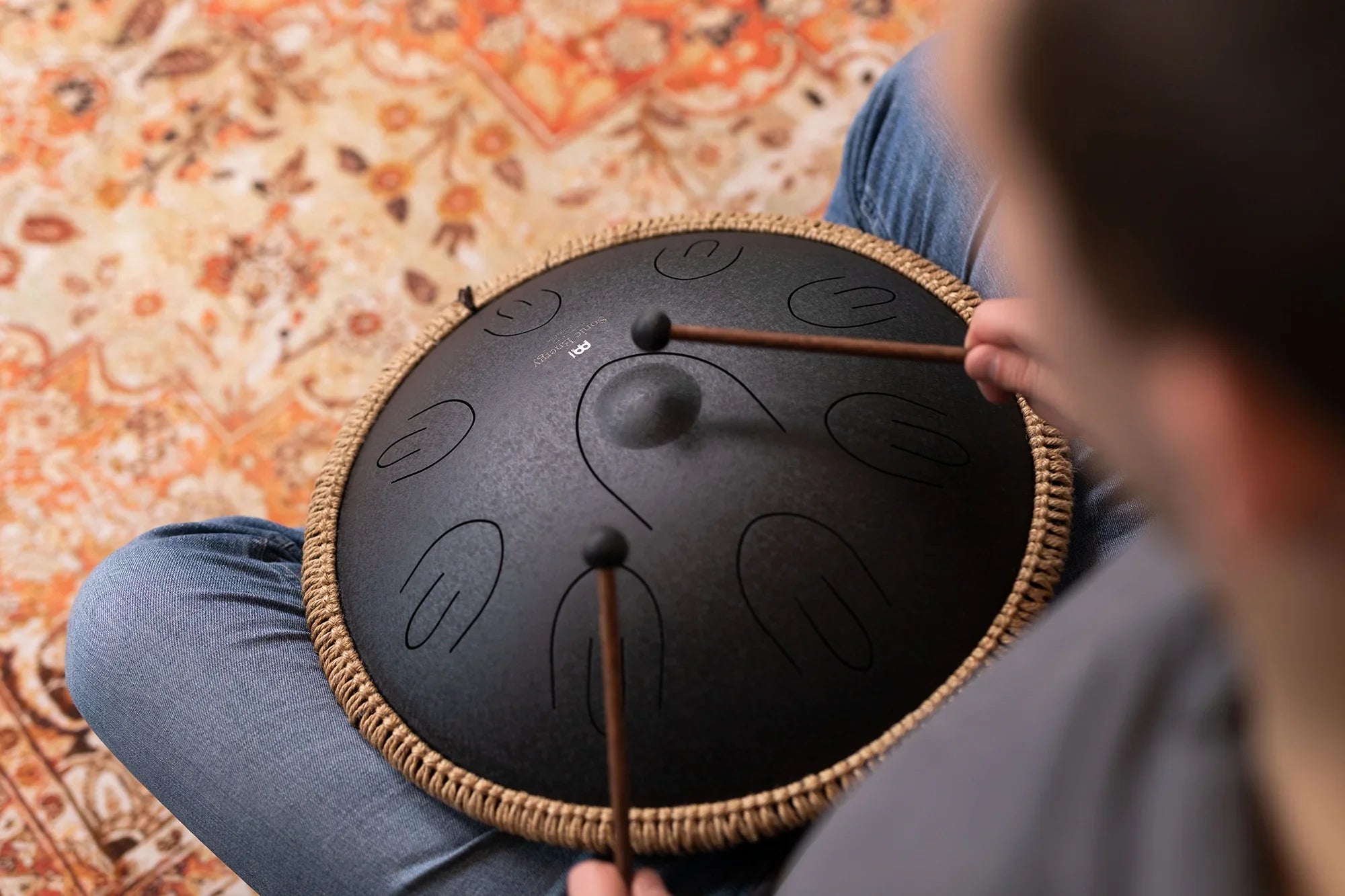 Person playing black tongue drum with mallets on patterned rug, sound healing instrument.
