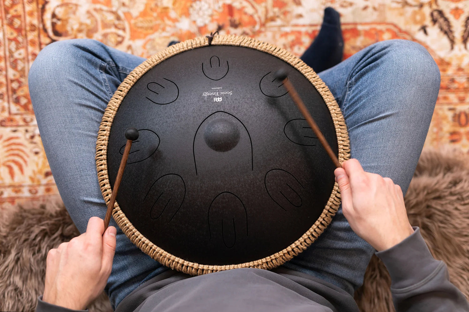 Person playing a black steel tongue drum with mallets, seated on a patterned rug