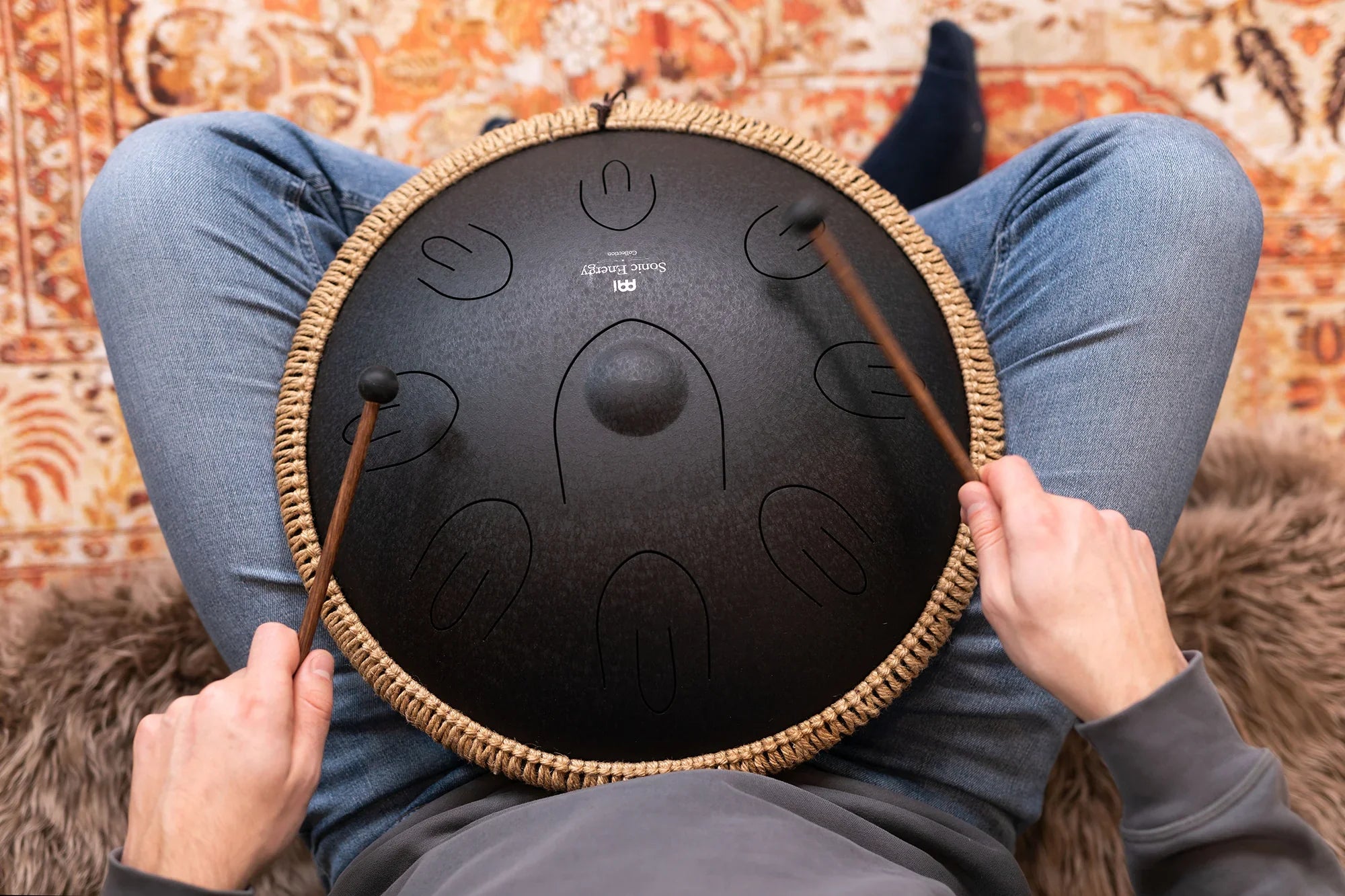 Person playing a black steel tongue drum with mallets, seated on a patterned rug