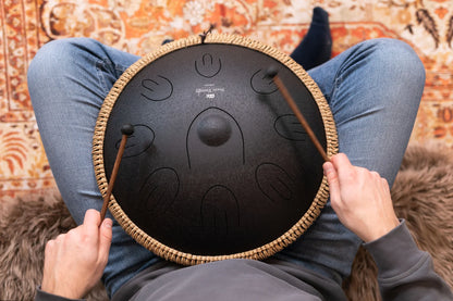 Person playing a black steel tongue drum with mallets, seated on a patterned rug