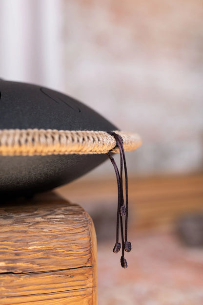 Close-up of a black steel tongue drum with rope trim, resting on a rustic wooden surface