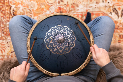 Person playing handpan tongue drum with mallets, close-up, on patterned rug background