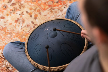 Person playing a steel tongue drum with mallets, seated on a patterned rug