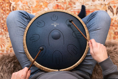 Person playing a steel tongue drum with mallets, sitting on the floor