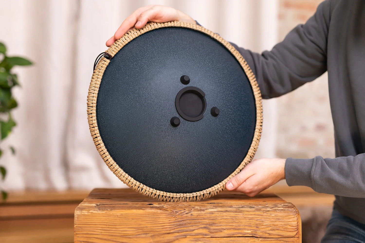 Hand holding a round handpan drum with woven rim, displayed on a wooden table indoors