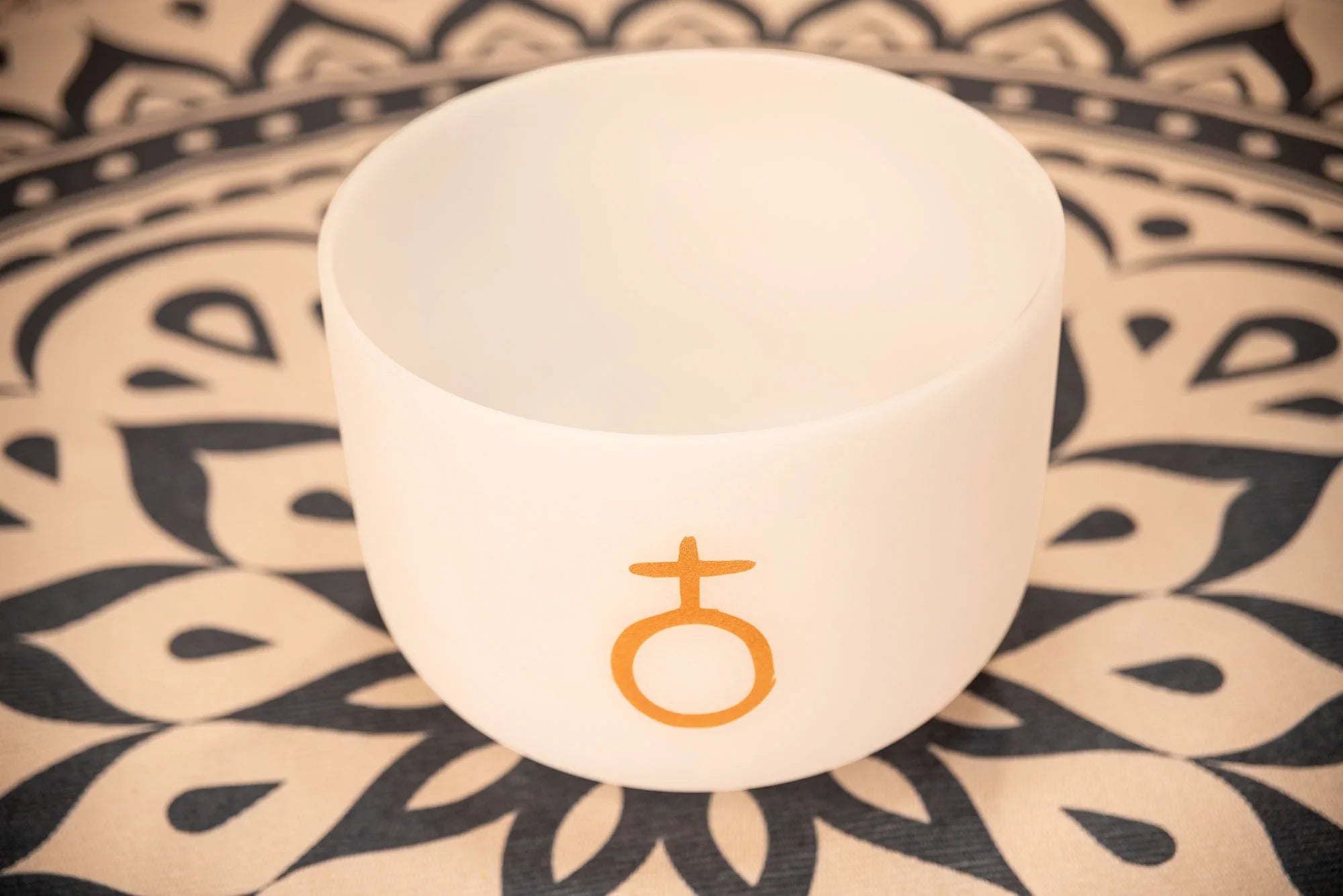 White crystal singing bowl with gold symbol on mandala patterned mat, sound healing instrument