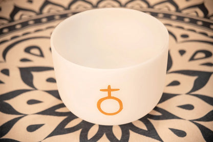 White crystal singing bowl with gold symbol on mandala patterned mat, sound healing instrument