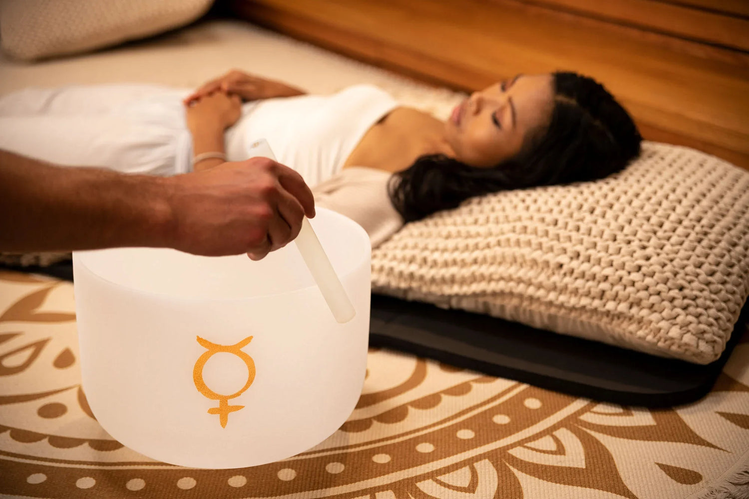Person playing crystal singing bowl during sound healing session with woman relaxing on mat