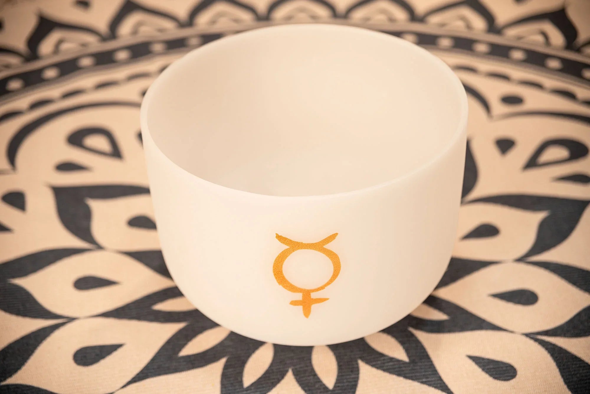 White crystal singing bowl with gold symbol on patterned mat, sound healing instrument