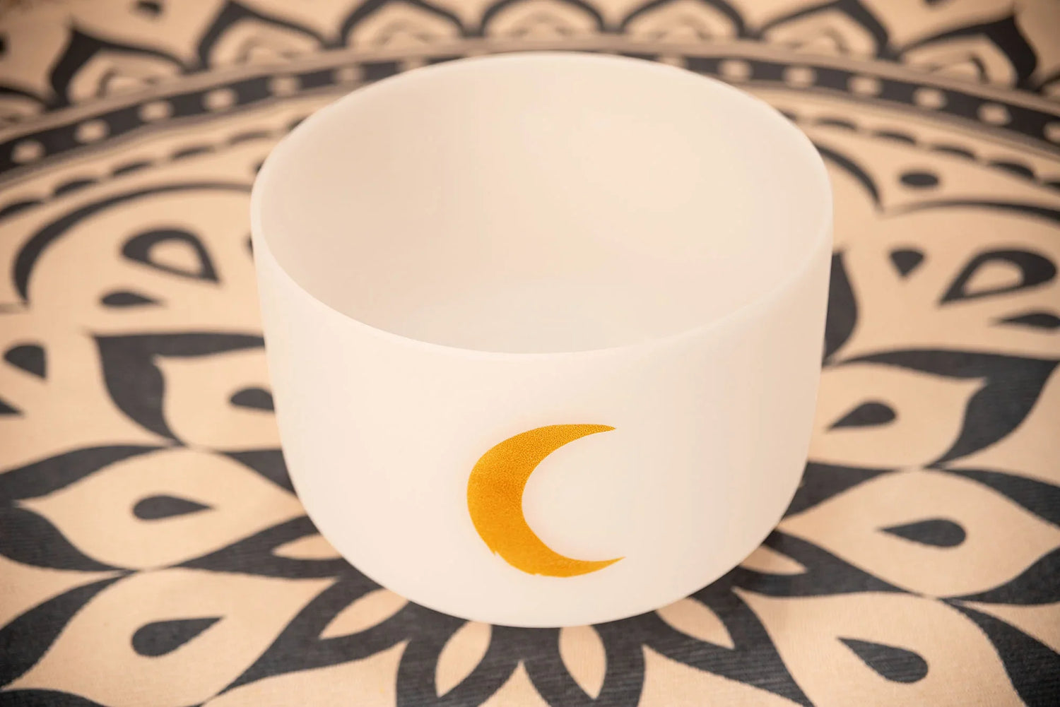 White crystal singing bowl with gold crescent moon, on a patterned mat, for sound healing