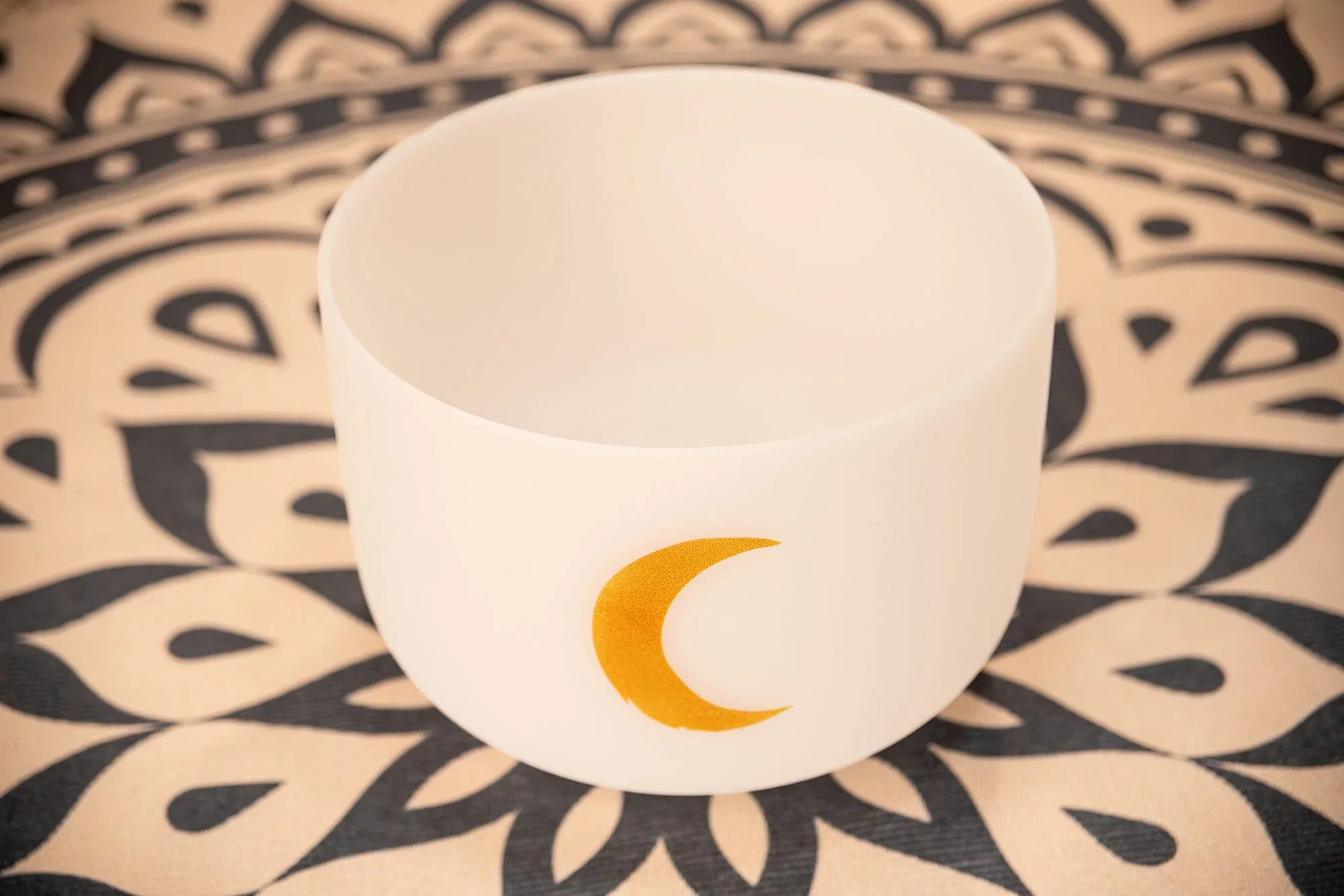 White crystal singing bowl with gold crescent moon, on a patterned mat, for sound healing