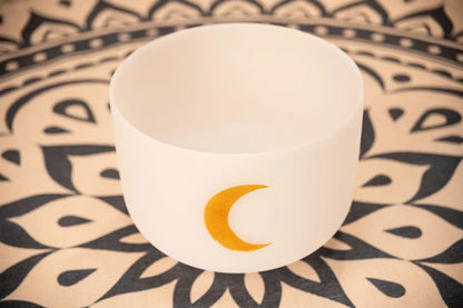 White crystal singing bowl with gold crescent moon, on a patterned mat, for sound healing