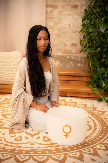 Woman meditating with white crystal singing bowl on patterned rug, sound healing setting
