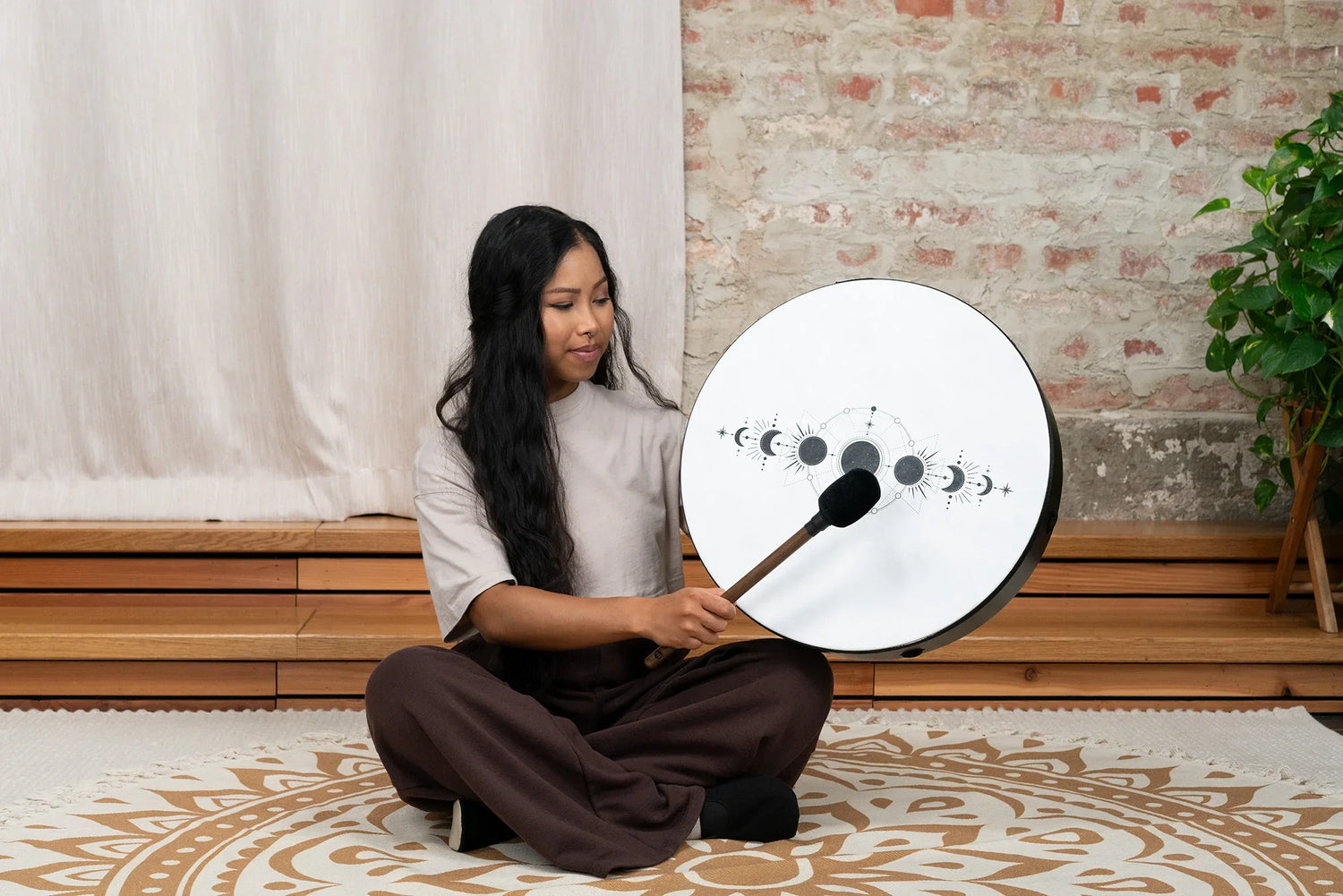 Woman sitting on mandala rug playing decorated frame drum for sound healing indoors