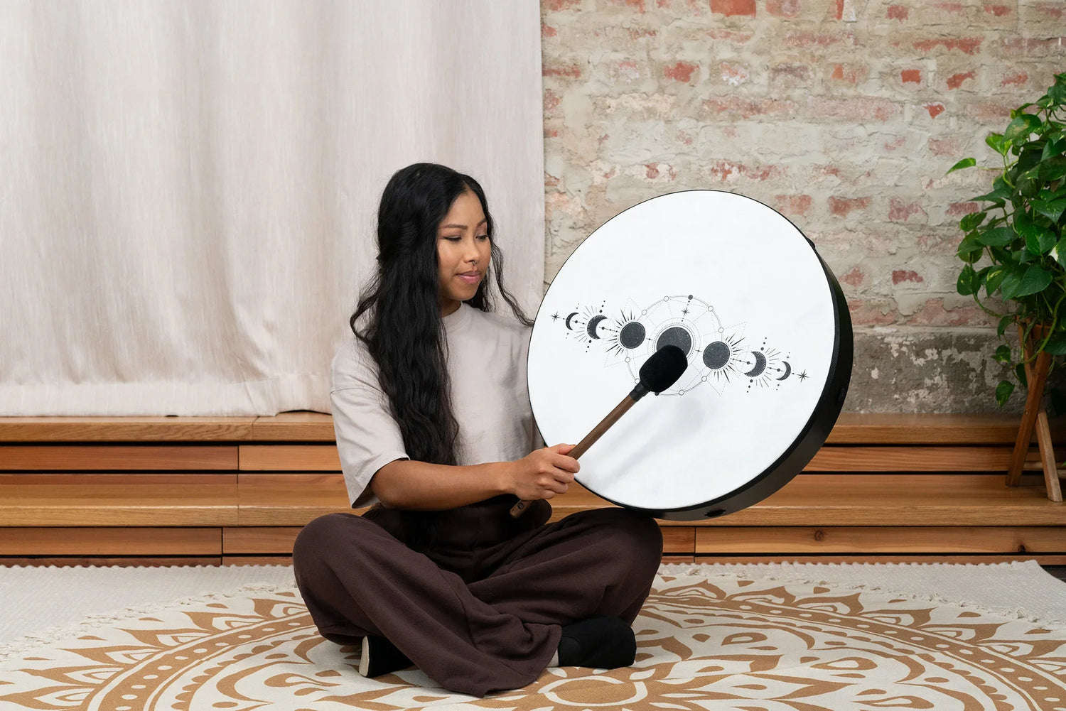 Person playing a hand drum with lunar phase design, seated on a mandala floor mat