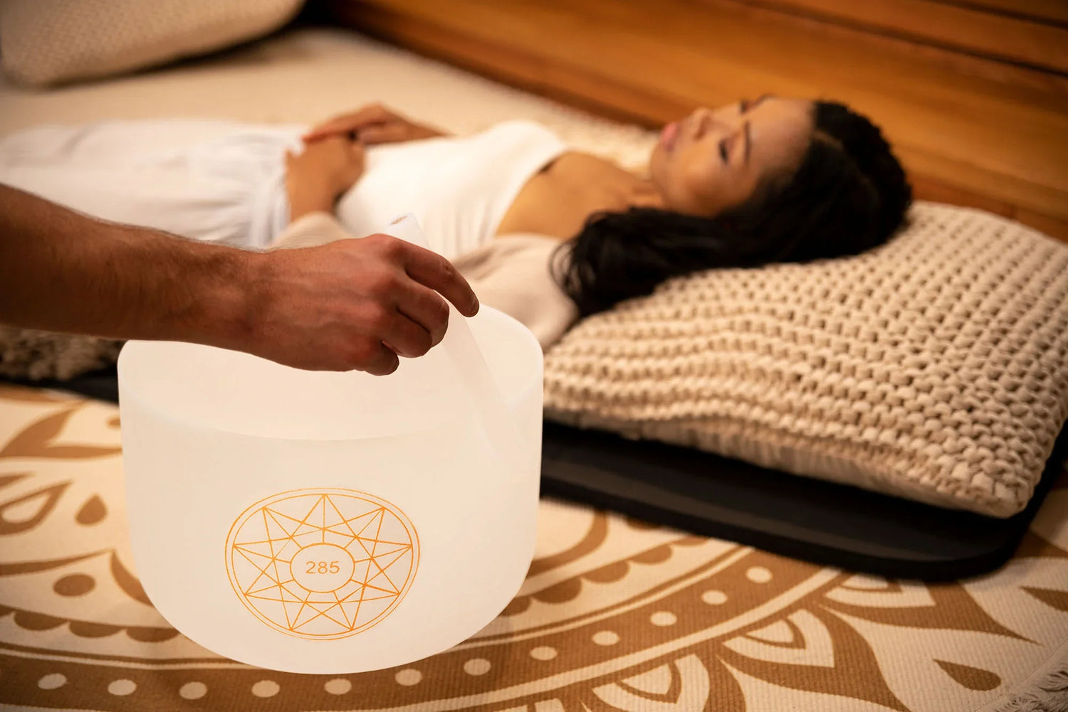 Person playing crystal singing bowl near woman lying on mat for sound healing session