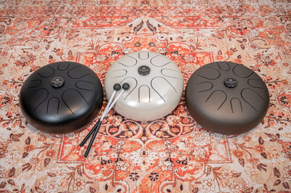Three steel tongue drums in black, white, and bronze on a patterned rug with mallets