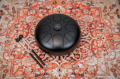 Black steel tongue drum with mallets on ornate patterned rug, sound healing instrument