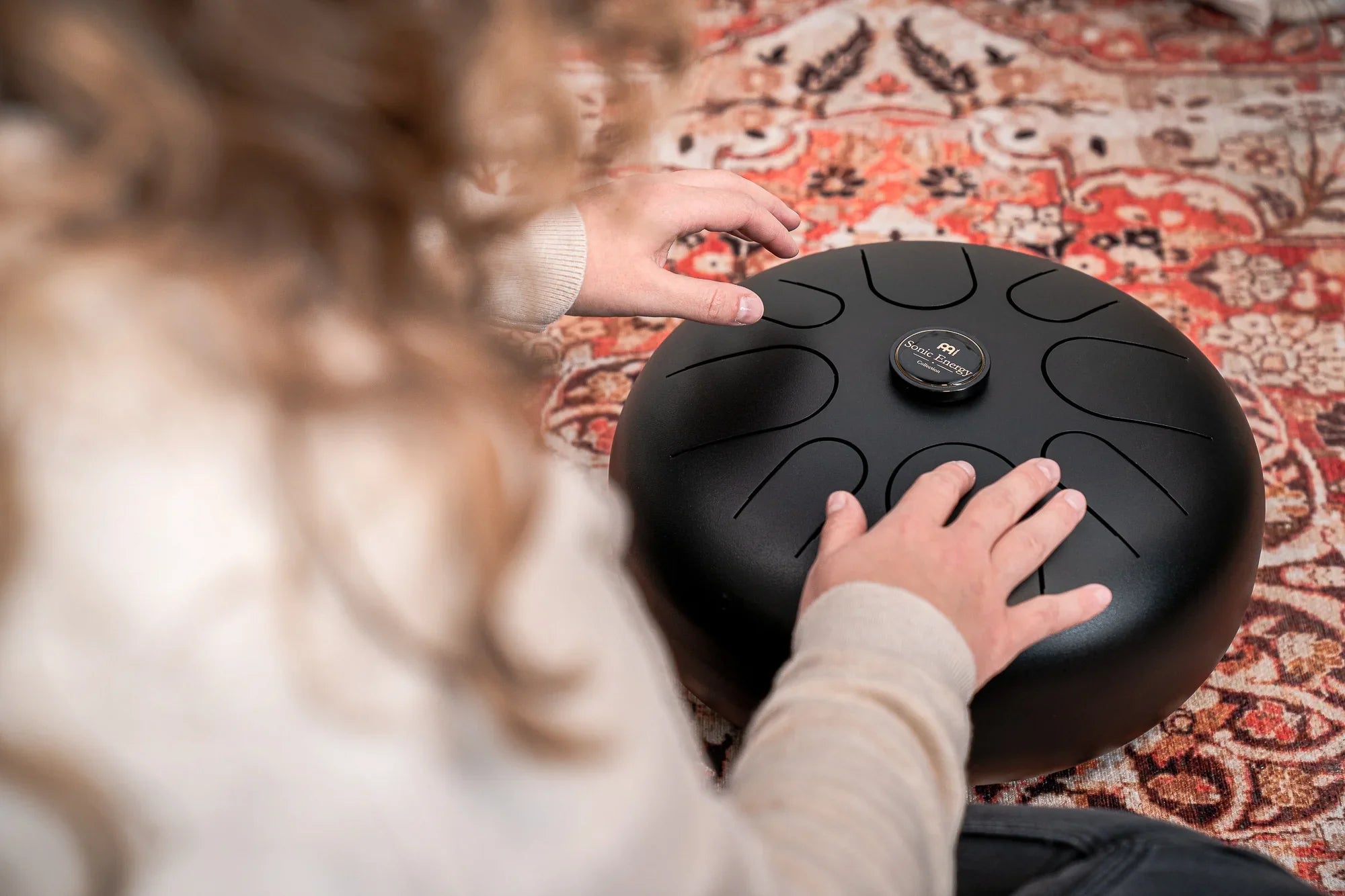 Hands playing a black tongue drum for sound healing on a patterned rug.