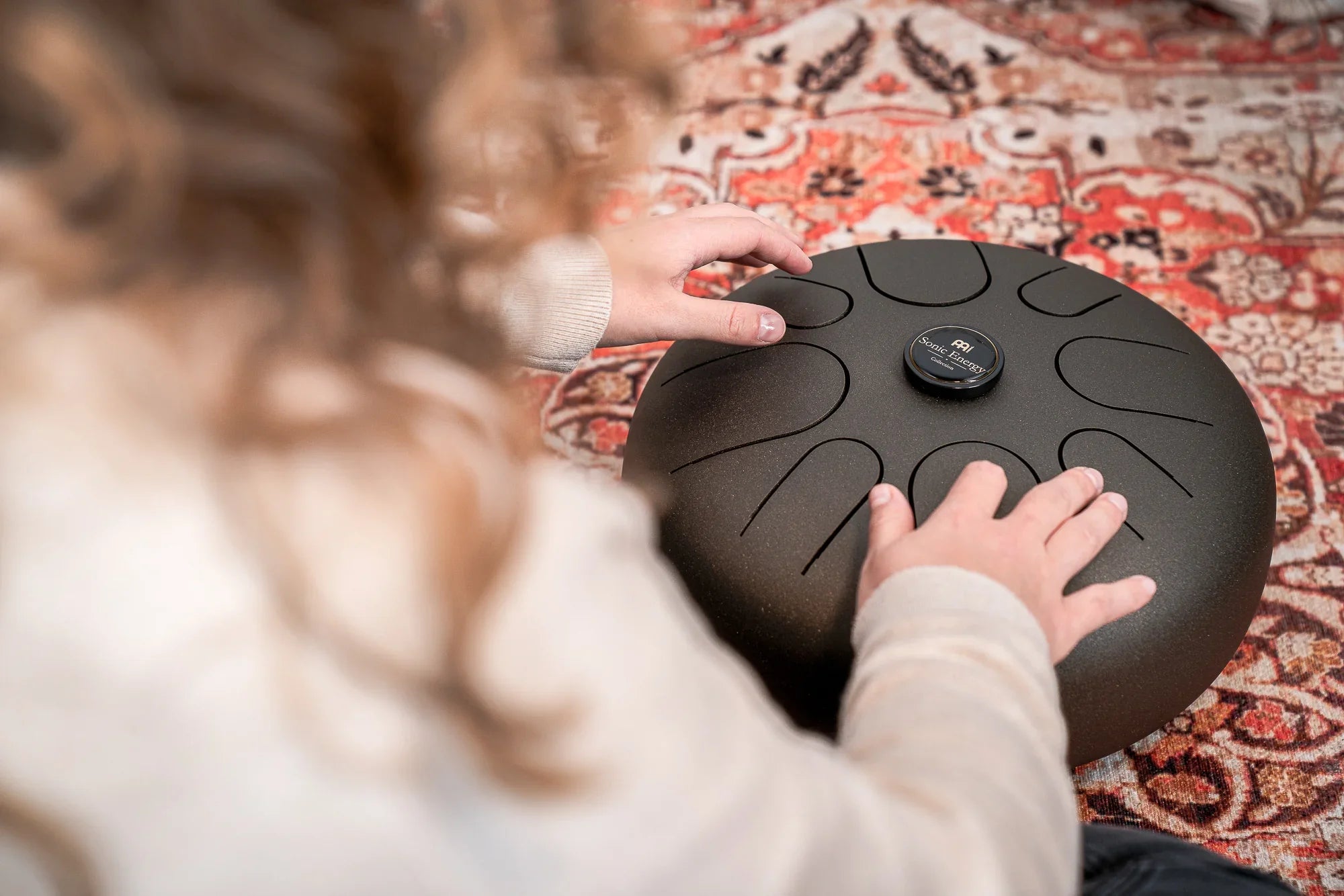 Person playing a black steel tongue drum on a patterned rug, sound healing instrument